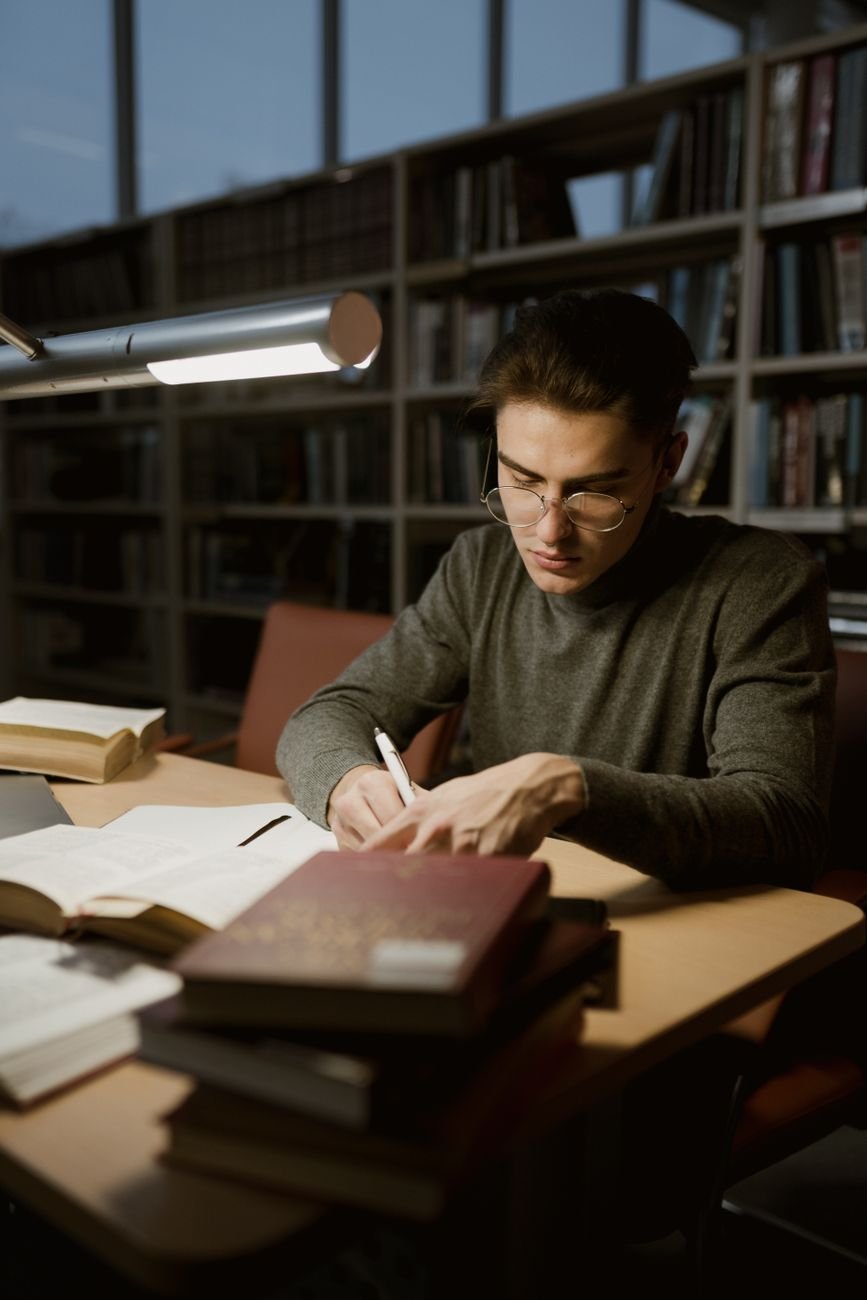A young man in a library studying with books and taking notes under a desk lamp.